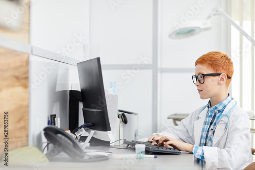Wallpaper Mural Side view portrait of cute red haired boy playing doctor sitting at desk in office and using computer, copy space Torontodigital.ca