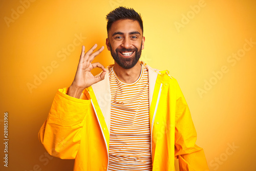 Young indian man wearing raincoat standing over isolated yellow background smiling positive doing ok sign with hand and fingers. Successful expression.