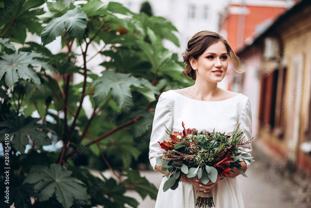 Portrait of a beautiful bride in a modest dress. Young brunette girl in a white dress with long sleeves. Cute bride with an autumn bouquet. Portrait of a woman outdoors on an autumn street.