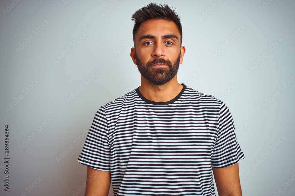 Young indian man wearing black striped t-shirt standing over isolated white background with serious expression on face. Simple and natural looking at the camera. Young indian man wearing black striped t-shirt standing over isolated white background with serious expression on face. Simple and natural looking at the camera.