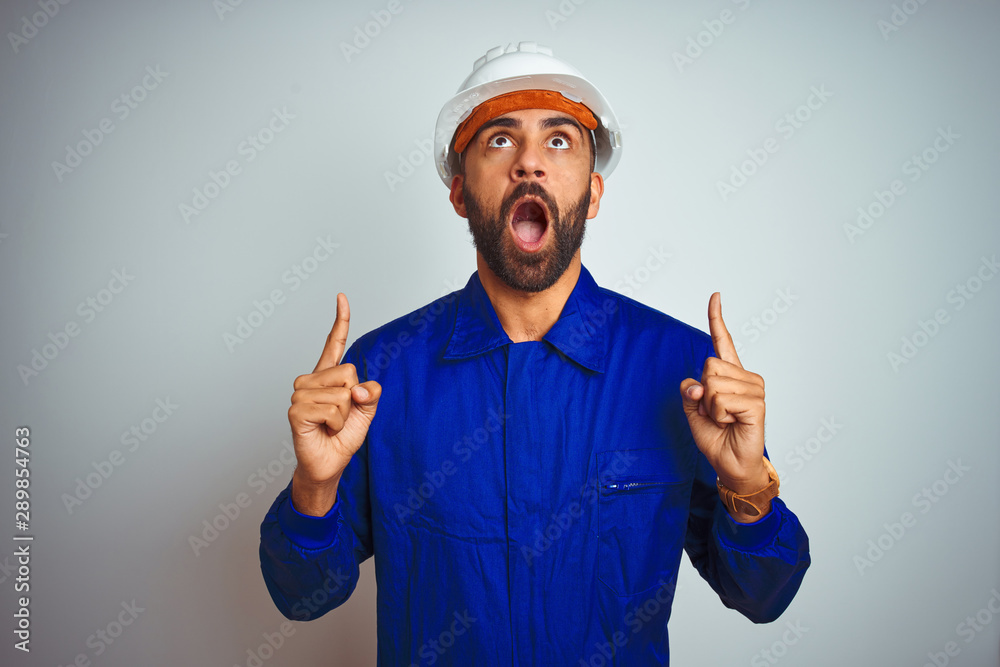 Handsome indian worker man wearing uniform and helmet over isolated white background amazed and surprised looking up and pointing with fingers and raised arms. Handsome indian worker man wearing uniform and helmet over isolated white background amazed and surprised looking up and pointing with fingers and raised arms.