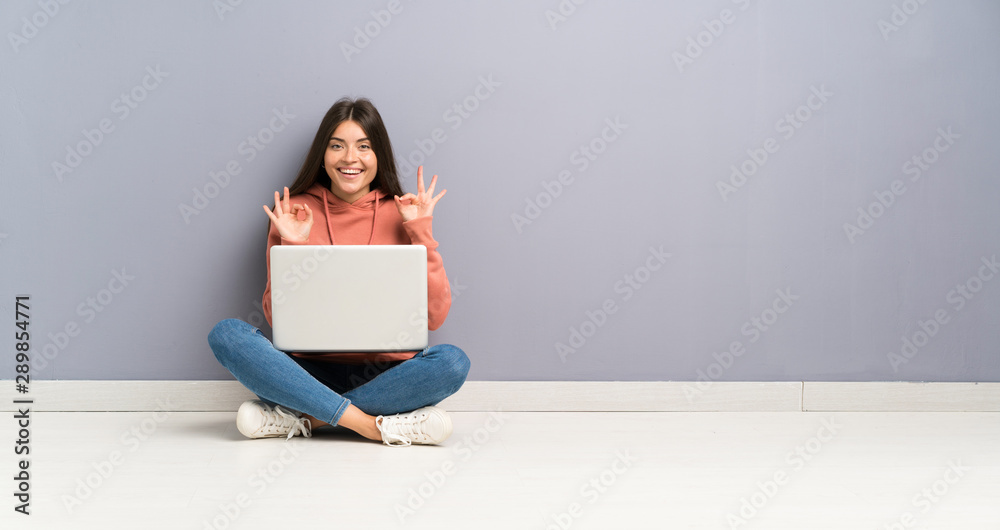 Young student girl with a laptop on the floor showing an ok sign with fingers Young student girl with a laptop on the floor showing an ok sign with fingers