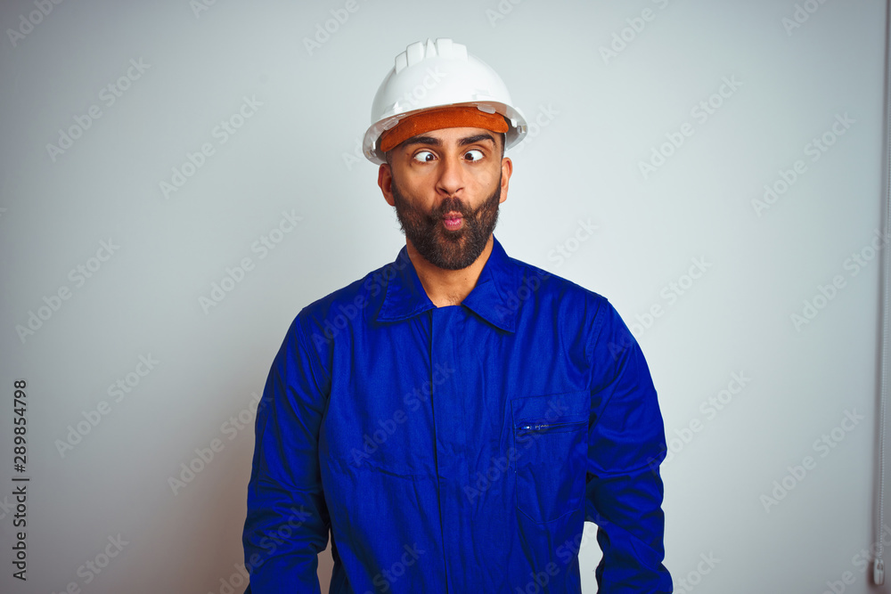 Handsome indian worker man wearing uniform and helmet over isolated white background making fish face with lips, crazy and comical gesture. Funny expression.