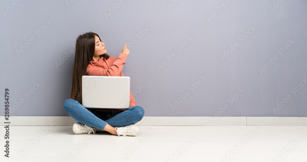 Young student girl with a laptop on the floor pointing back with the index finger Young student girl with a laptop on the floor pointing back with the index finger
