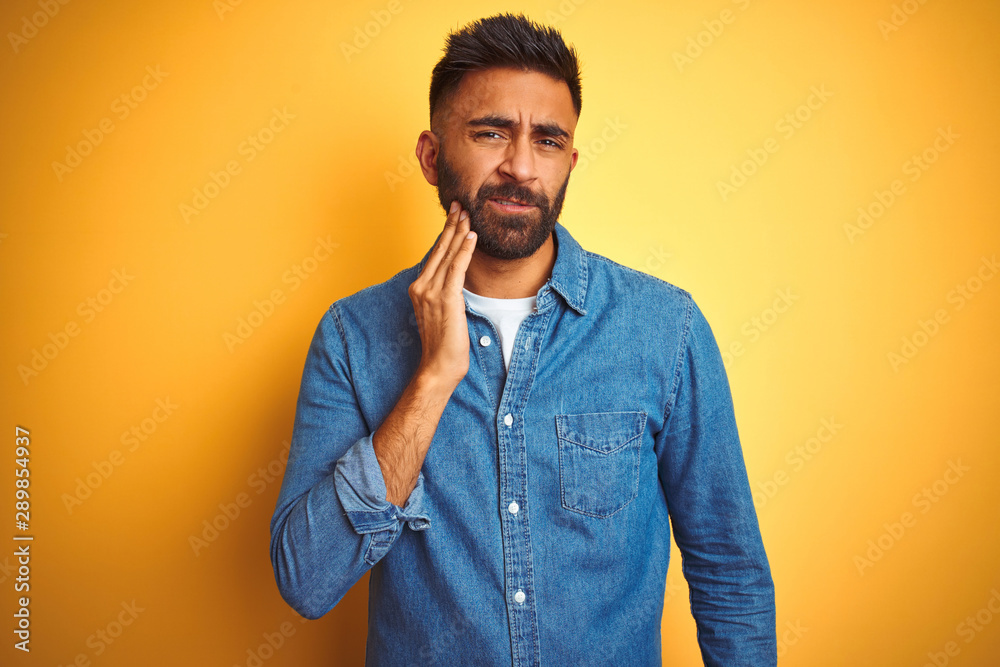 Young indian man wearing denim shirt standing over isolated yellow background touching mouth with hand with painful expression because of toothache or dental illness on teeth. Dentist concept. Young indian man wearing denim shirt standing over isolated yellow background touching mouth with hand with painful expression because of toothache or dental illness on teeth. Dentist concept.