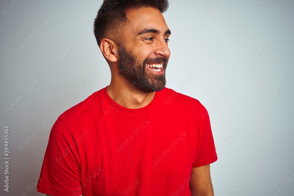 Young indian man wearing red t-shirt over isolated white background looking away to side with smile on face, natural expression. Laughing confident.