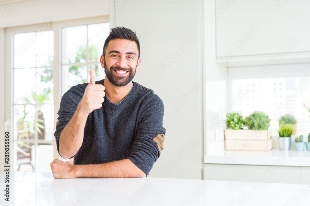 Handsome hispanic man wearing casual sweater at home doing happy thumbs up gesture with hand. Approving expression looking at the camera showing success.