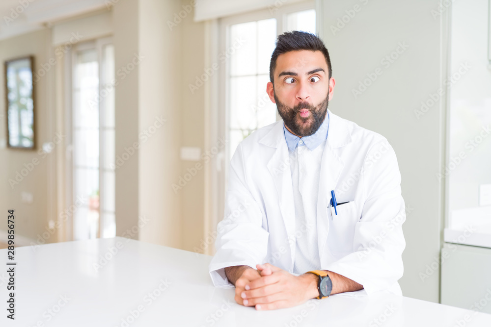Handsome hispanic doctor or therapist man wearing medical coat at the clinic making fish face with lips, crazy and comical gesture. Funny expression.