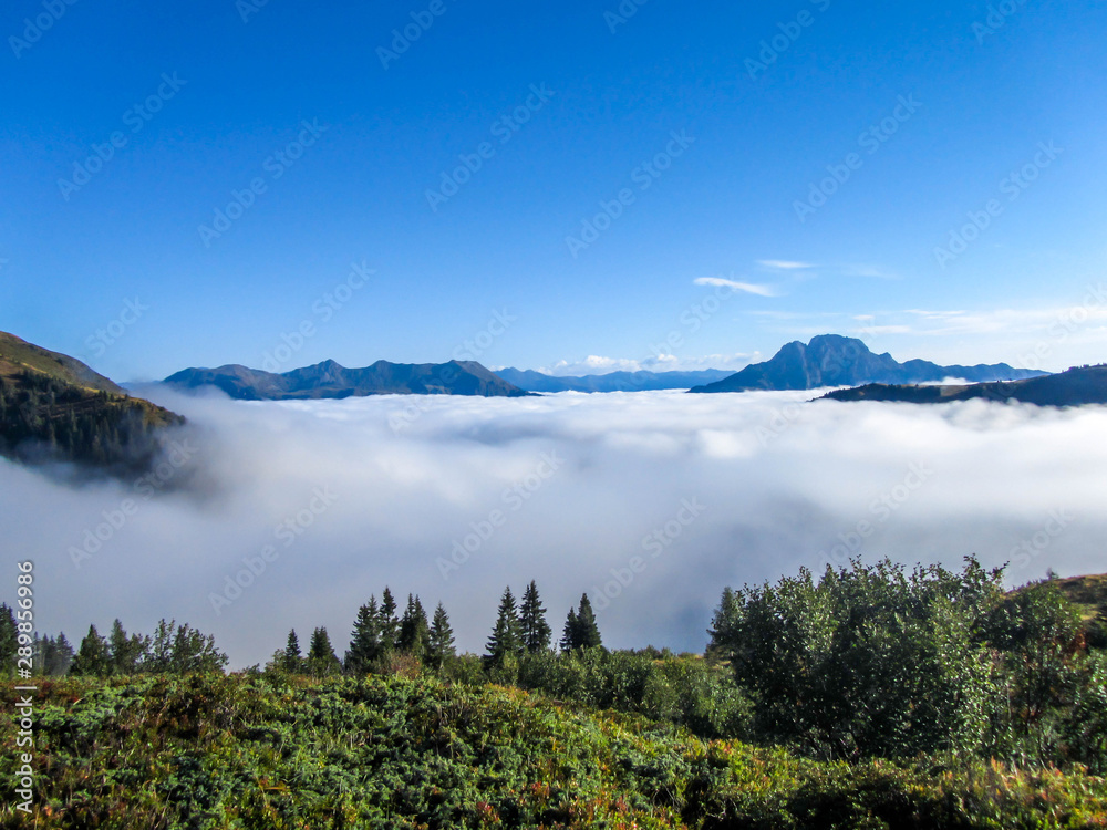 A panoramic view on Alpine valley. The valley is shrouded in dense fog, only tall mountain peaks are visible. Hiking trails in the high mountain. Endless chains of mountains visible. Magical mist.