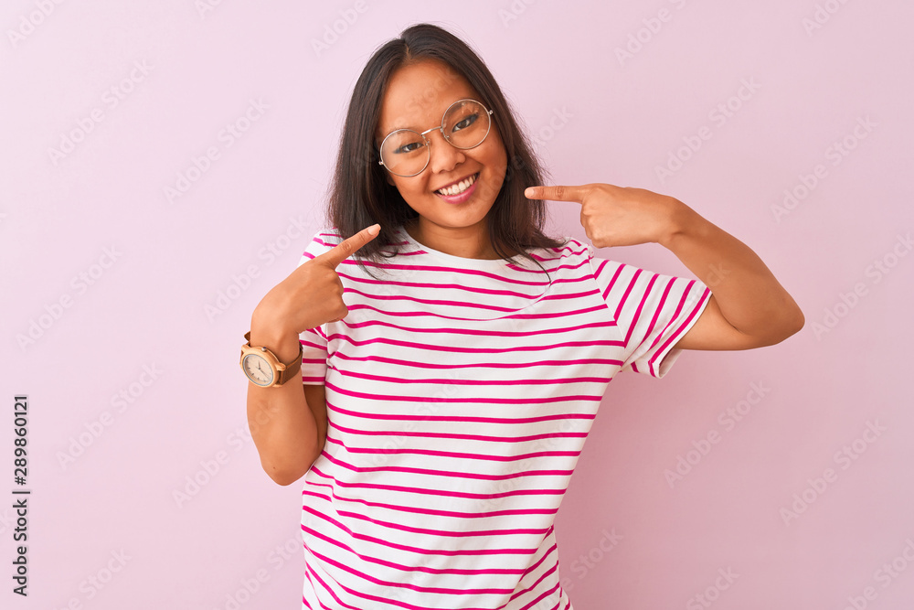 Young chinese woman wearing striped t-shirt and glasses over isolated pink background smiling cheerful showing and pointing with fingers teeth and mouth. Dental health concept.
