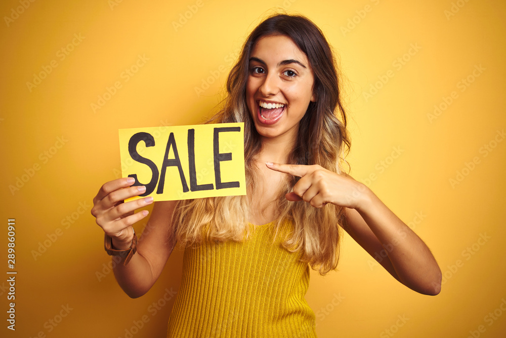 Young beautiful woman holding sale poster over yellow isolated background very happy pointing with hand and finger Young beautiful woman holding sale poster over yellow isolated background very happy pointing with hand and finger