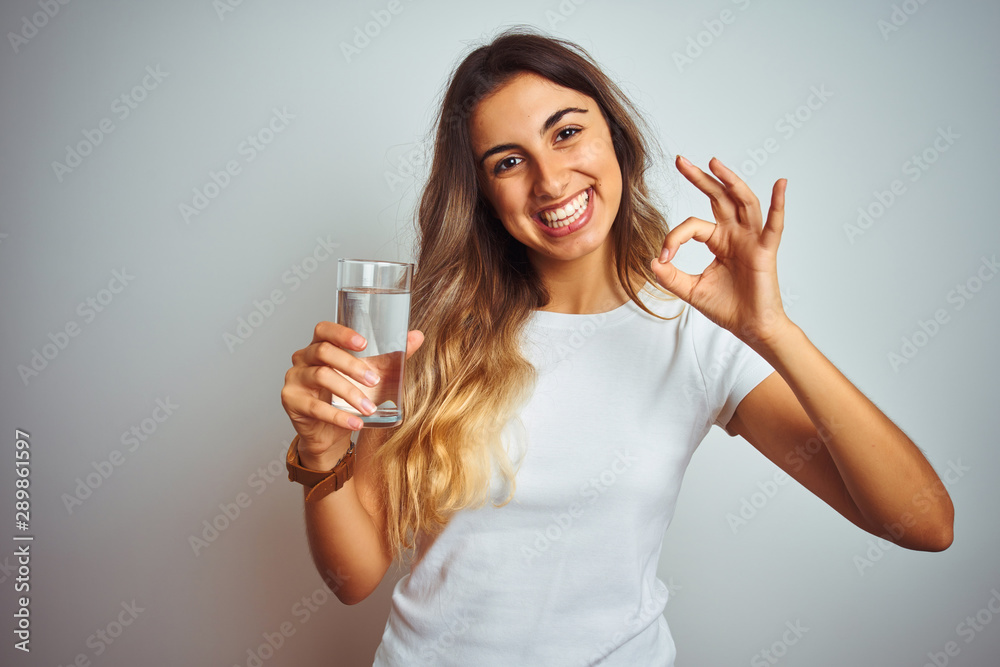 Young beautiful woman drinking a glass of water over white isolated background doing ok sign with fingers, excellent symbol Young beautiful woman drinking a glass of water over white isolated background doing ok sign with fingers, excellent symbol