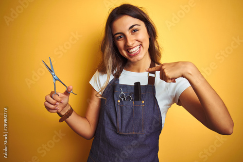 Young beautiful hairdresser woman holding scissors over yellow isolated background with surprise face pointing finger to himself