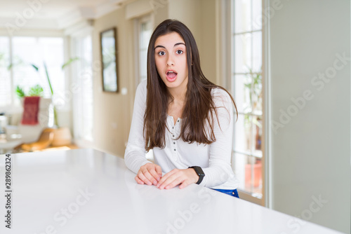 Beautiful young woman sitting on white table at home afraid and shocked with surprise expression, fear and excited face. © Krakenimages.com