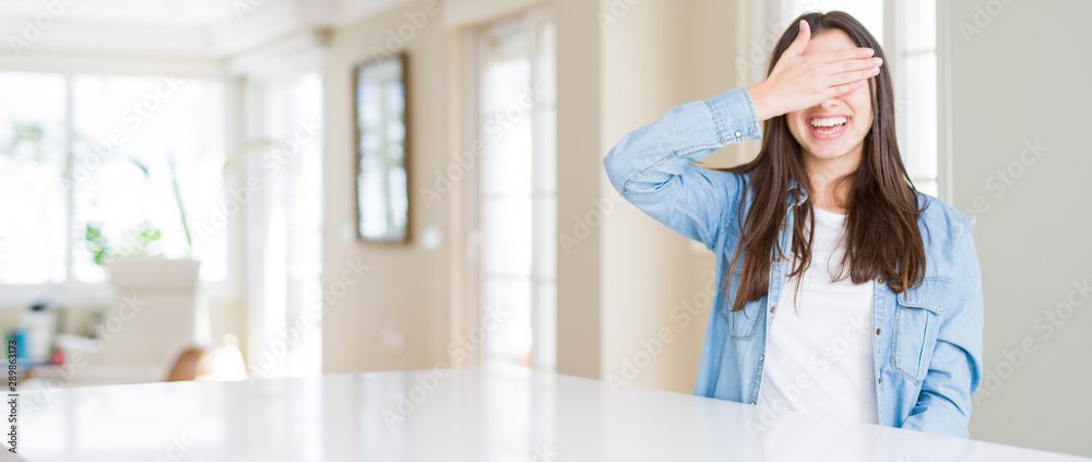 Wide angle picture of beautiful young woman sitting on white table at home smiling and laughing with hand on face covering eyes for surprise. Blind concept. Wide angle picture of beautiful young woman sitting on white table at home smiling and laughing with hand on face covering eyes for surprise. Blind concept.