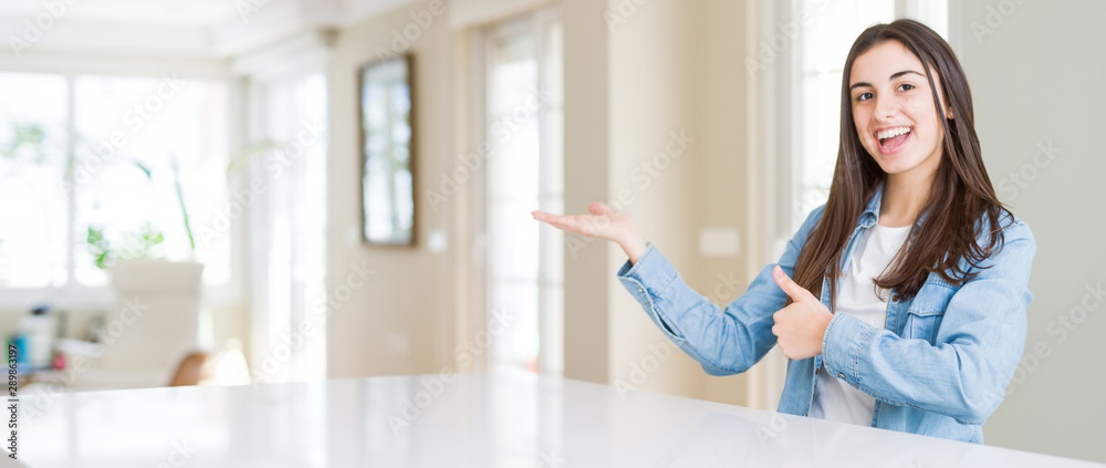 Wide angle picture of beautiful young woman sitting on white table at home Showing palm hand and doing ok gesture with thumbs up, smiling happy and cheerful