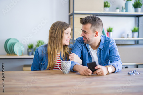 Young couple relaxing drinking a cup of coffee and using smartphone, sitting at new home with cardboard boxes behind them