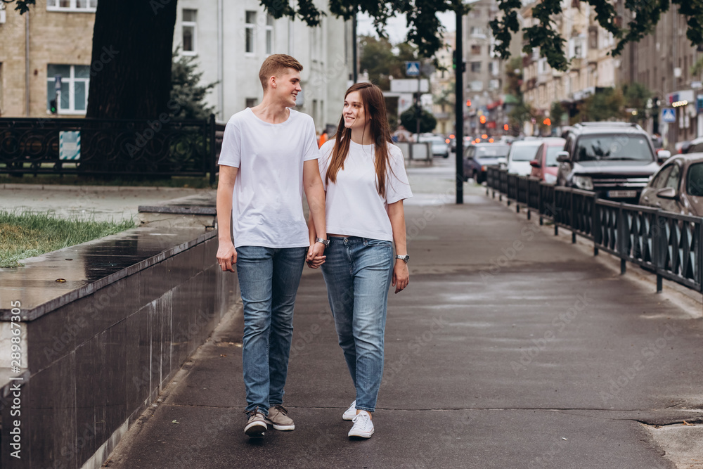 young couple walks the streets of the city and hold hands. guy and girl in white t-shirts and jeans outdoors. Teenagers walk in the autumn city. Happy young couple. young couple walks the streets of the city and hold hands. guy and girl in white t-shirts and jeans outdoors. Teenagers walk in the autumn city. Happy young couple.