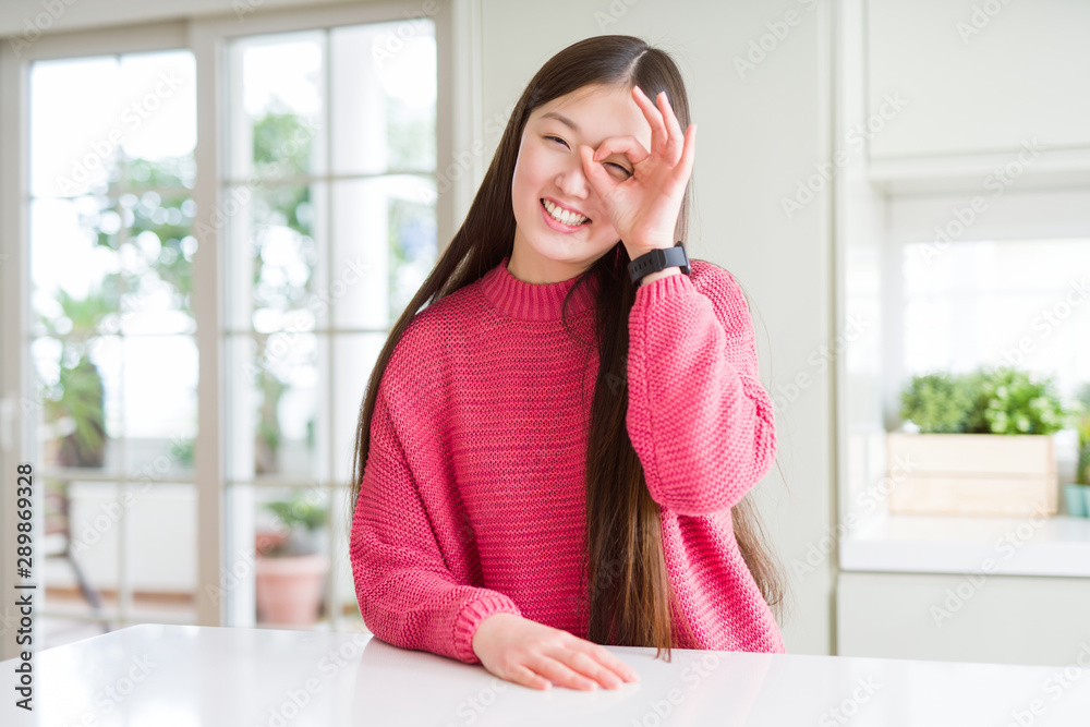 Beautiful Asian woman wearing pink sweater on white table doing ok gesture with hand smiling, eye looking through fingers with happy face. Beautiful Asian woman wearing pink sweater on white table doing ok gesture with hand smiling, eye looking through fingers with happy face.