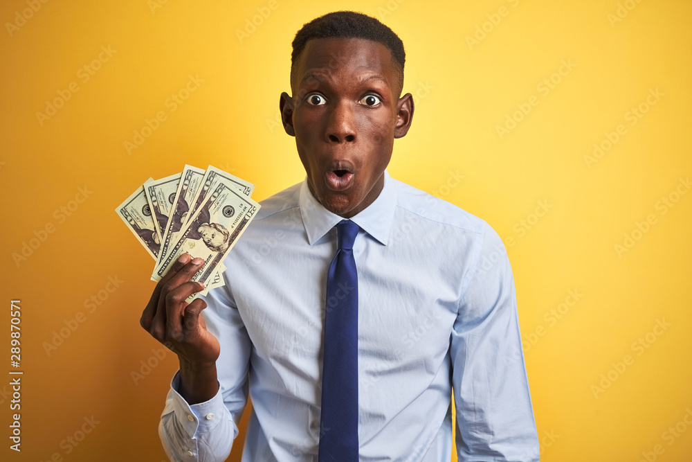 African american businessman holding dollars standing over isolated yellow background scared in shock with a surprise face, afraid and excited with fear expression African american businessman holding dollars standing over isolated yellow background scared in shock with a surprise face, afraid and excited with fear expression
