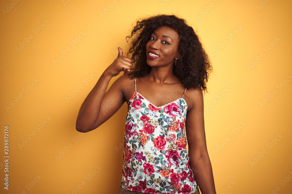 African american woman wearing floral summer t-shirt over isolated yellow background smiling doing phone gesture with hand and fingers like talking on the telephone. Communicating concepts.