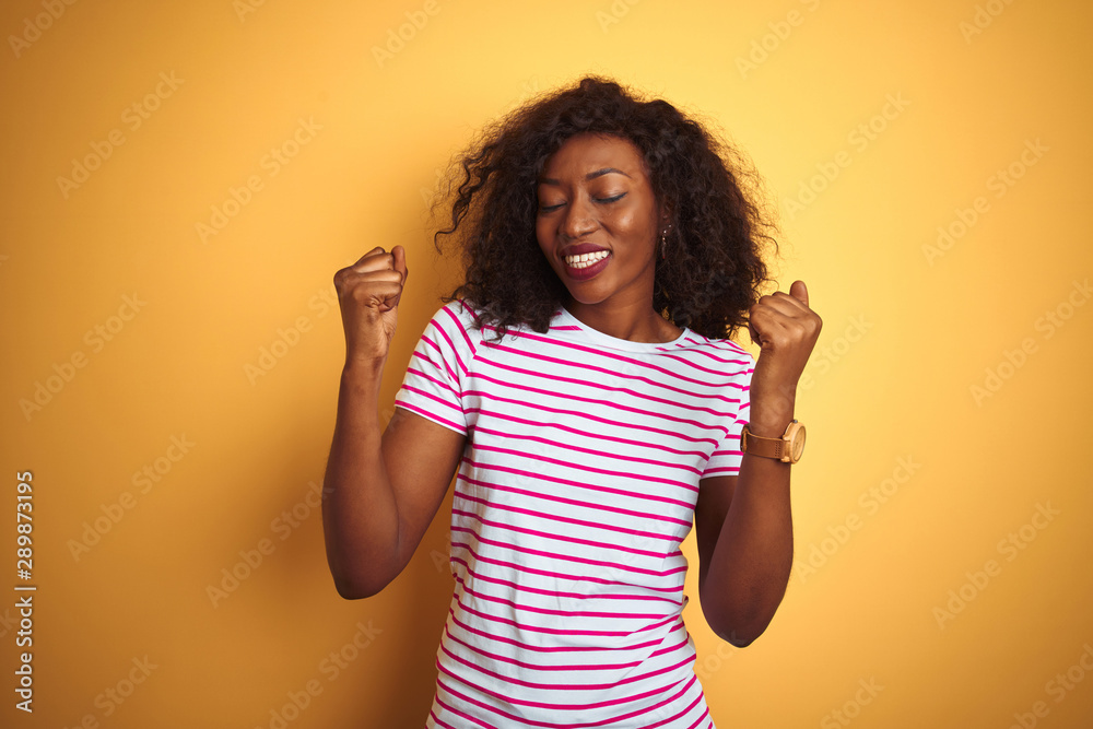Young african american woman wearing striped t-shirt over isolated yellow background very happy and excited doing winner gesture with arms raised, smiling and screaming for success. Young african american woman wearing striped t-shirt over isolated yellow background very happy and excited doing winner gesture with arms raised, smiling and screaming for success.