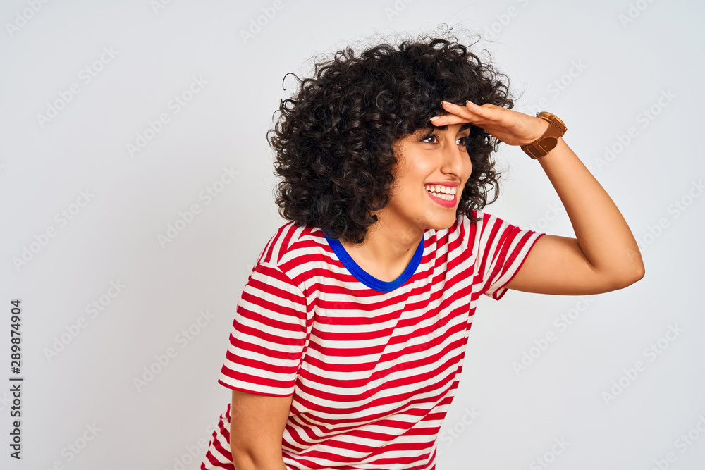 Young arab woman with curly hair wearing striped t-shirt over isolated white background very happy and smiling looking far away with hand over head. Searching concept. Young arab woman with curly hair wearing striped t-shirt over isolated white background very happy and smiling looking far away with hand over head. Searching concept.