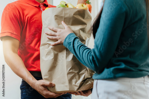 smart food delivery service man in red uniform handing fresh food to recipient and young woman customer receiving order from courier at home, express delivery, food delivery, online shopping concept