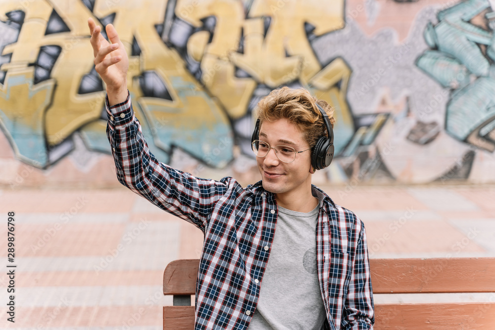 Obraz premium young man listening to music is waving hand and sitting on a bench