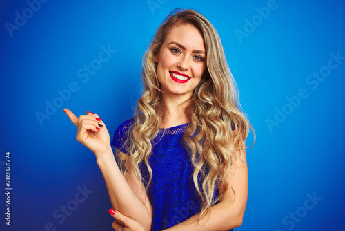 Young beautiful woman standing over blue isolated background with a big smile on face, pointing with hand and finger to the side looking at the camera.