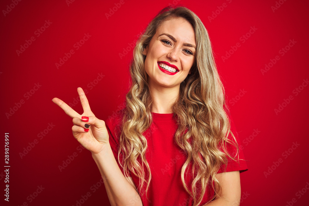 Young beautiful woman wearing basic t-shirt standing over red isolated background smiling with happy face winking at the camera doing victory sign. Number two. Young beautiful woman wearing basic t-shirt standing over red isolated background smiling with happy face winking at the camera doing victory sign. Number two.