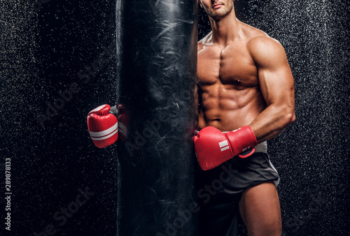 Beautiful muscular fighter with naked torso is standing at dark room with water drops on the wall. © Fxquadro