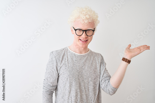Young albino blond man wearing striped t-shirt and glasses over isolated white background smiling cheerful presenting and pointing with palm of hand looking at the camera.