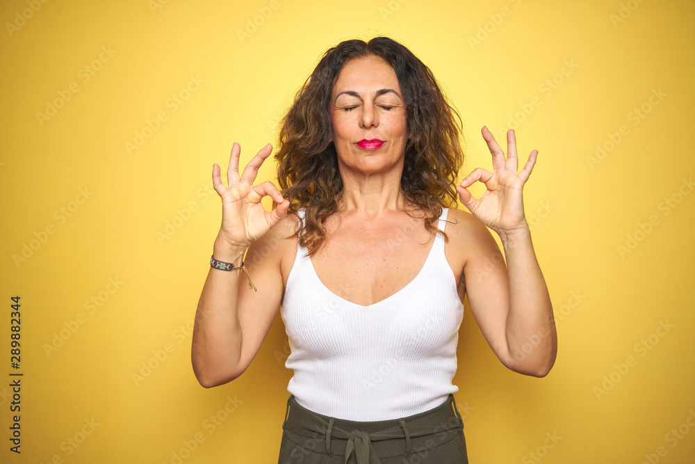 Middle age senior woman with curly hair standing over yellow isolated background relax and smiling with eyes closed doing meditation gesture with fingers. Yoga concept. Middle age senior woman with curly hair standing over yellow isolated background relax and smiling with eyes closed doing meditation gesture with fingers. Yoga concept.