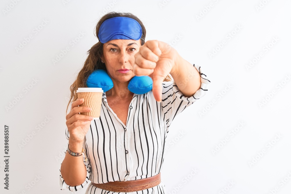 Tourist woman wearing neckpillow sleep mask drinking coffee over isolated white background with angry face, negative sign showing dislike with thumbs down, rejection concept Tourist woman wearing neckpillow sleep mask drinking coffee over isolated white background with angry face, negative sign showing dislike with thumbs down, rejection concept