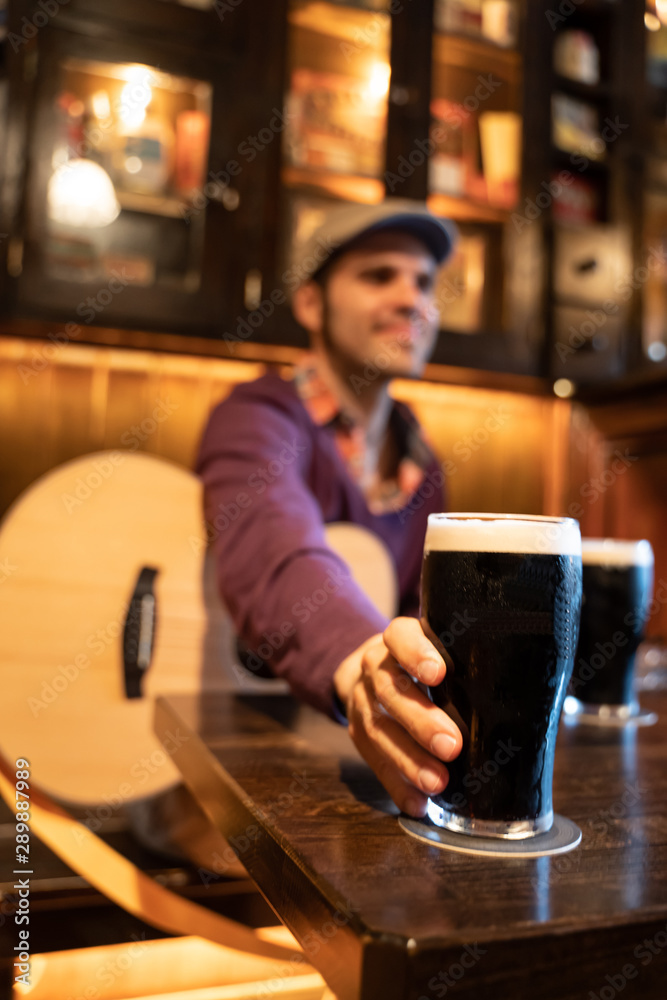 Man sitting on a table with a acoustic guitar taking a dark beer glass
