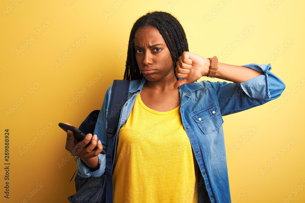 African american student woman using smartphone standing over isolated yellow background with angry face, negative sign showing dislike with thumbs down, rejection concept African american student woman using smartphone standing over isolated yellow background with angry face, negative sign showing dislike with thumbs down, rejection concept