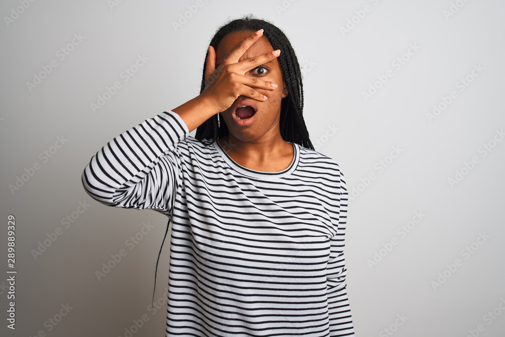 Young african american woman wearing striped t-shirt standing over isolated white background peeking in shock covering face and eyes with hand, looking through fingers with embarrassed expression. Young african american woman wearing striped t-shirt standing over isolated white background peeking in shock covering face and eyes with hand, looking through fingers with embarrassed expression.