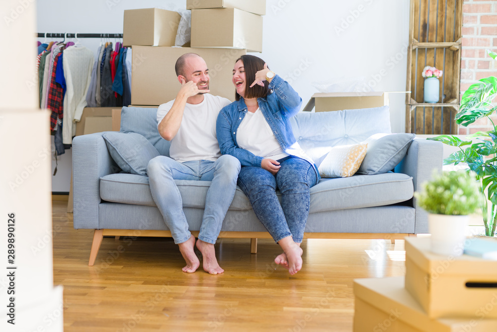 Young couple sitting on the sofa arround cardboard boxes moving to a new house smiling doing phone gesture with hand and fingers like talking on the telephone. Communicating concepts. Young couple sitting on the sofa arround cardboard boxes moving to a new house smiling doing phone gesture with hand and fingers like talking on the telephone. Communicating concepts.