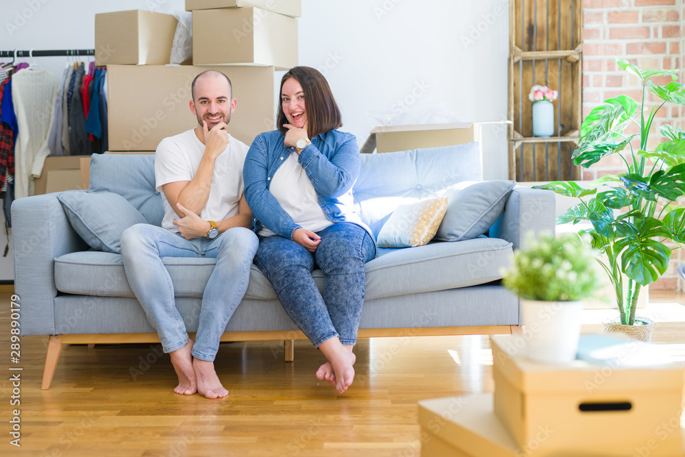 Young couple sitting on the sofa arround cardboard boxes moving to a new house looking confident at the camera smiling with crossed arms and hand raised on chin. Thinking positive. Young couple sitting on the sofa arround cardboard boxes moving to a new house looking confident at the camera smiling with crossed arms and hand raised on chin. Thinking positive.