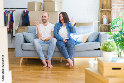 Young couple sitting on the sofa arround cardboard boxes moving to a new house smiling with happy face winking at the camera doing victory sign. Number two.