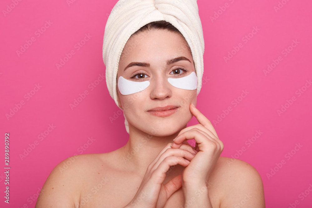 Close up portrait of beautiful young woman with collagen pads under her eyes, stands wearing white towel on head, charming gentle female keeps hands near her face. Spa, eye skin care concept. Close up portrait of beautiful young woman with collagen pads under her eyes, stands wearing white towel on head, charming gentle female keeps hands near her face. Spa, eye skin care concept.