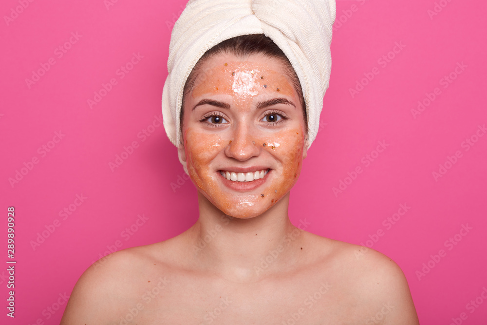 Horizontalshot of beautiful girl wrapped hair with white towel, skin covered by mask. Model with mask on face, posing with bare shoulders, standig indoor against pink studio wall. Beauty concept. Horizontalshot of beautiful girl wrapped hair with white towel, skin covered by mask. Model with mask on face, posing with bare shoulders, standig indoor against pink studio wall. Beauty concept.