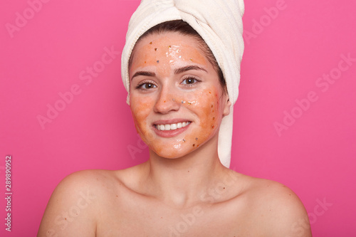 Horizontal shot of young woman with moisturizer mask on her face. Photo of smiling female receiving spa treatments, charming girl posing with bared shoulders and white tpowel on head. Beauty concept.