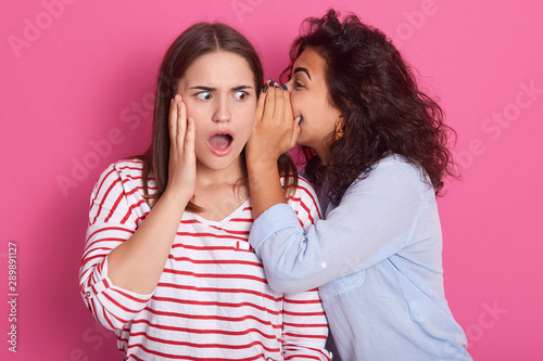 Horizontal shot of young woman whispers to her mate shocking news, brunette woman dresses striped casul shirt posing with open mouth, her friend with curly hair telling something interesting.