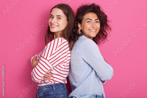 Profile of two attractive adult girls standing back to back, wearing casual clothing, looking smiling directly at camera, posing isolated over pink studio, females expressing happyness and joy. photo