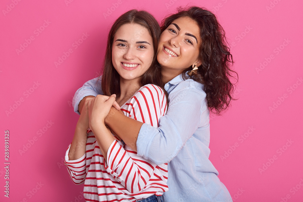 Smiling women embracing and looking directly at camera. Lesbian couple or women friendship concept. Isolated front view over pink background. Ladies wearing stylish shirt, expressing hapyness.