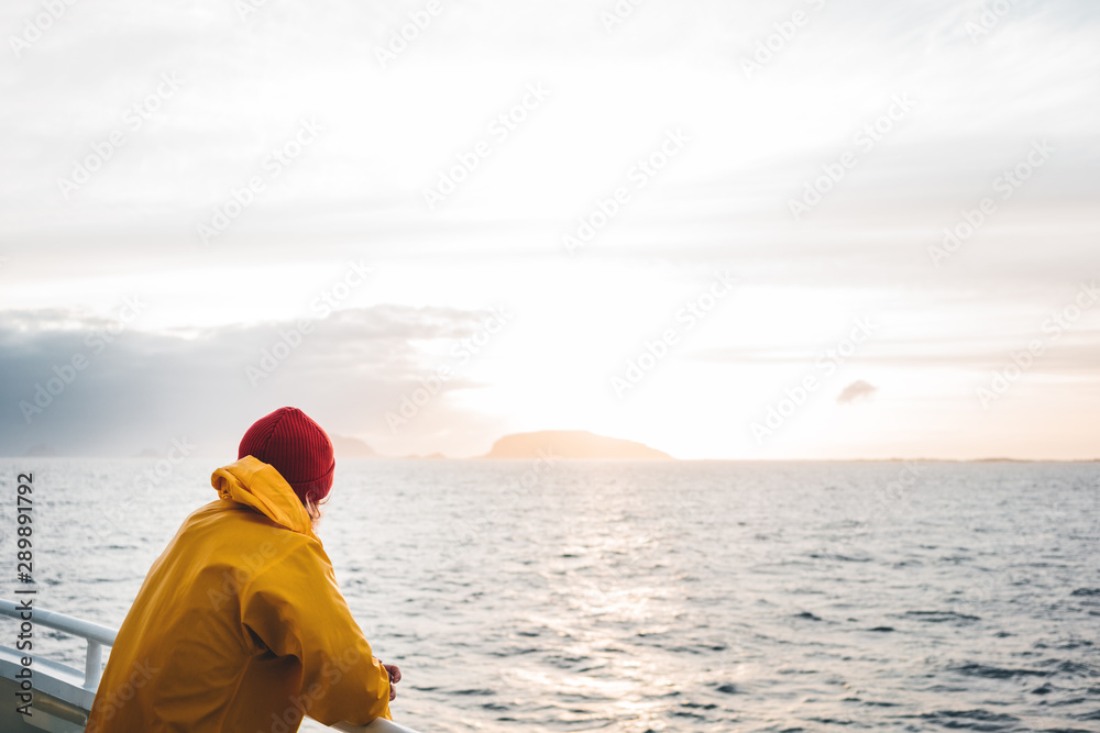 Fototapeta premium Hipster traveler wearing red hat and yellow raincoat looking away at cloudy mountain and sunset sea. Alone man traveling at scandinavian authentic ocean landscape by ship