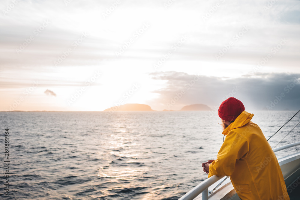 Hipster traveler wearing red hat and yellow raincoat looking away at cloudy mountain and sunset sea. Alone man traveling  at scandinavian authentic ocean landscape by ship