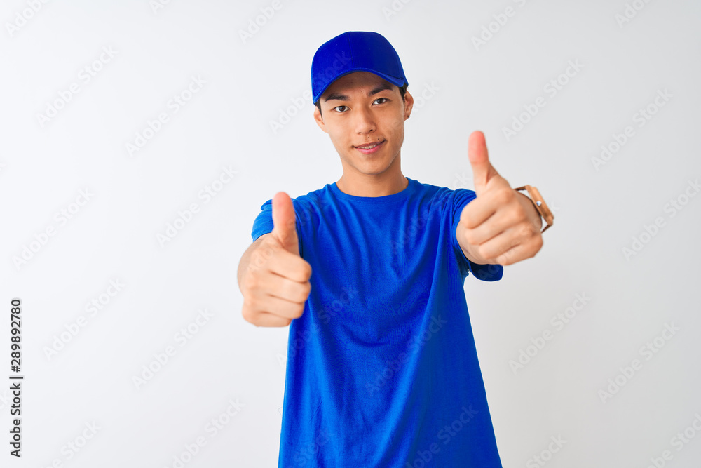 Chinese deliveryman wearing blue t-shirt and cap standing over isolated white background approving doing positive gesture with hand, thumbs up smiling and happy for success. Winner gesture.